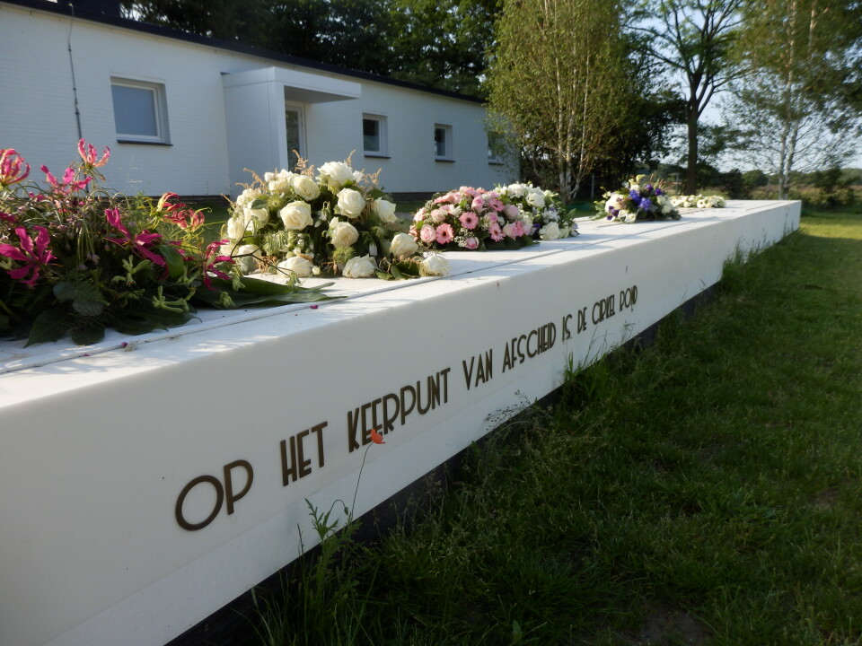 Bloemstukken op een witte muur met opschrift bij Crematorium Zuiderhof in Hilversum, omgeven door groen.