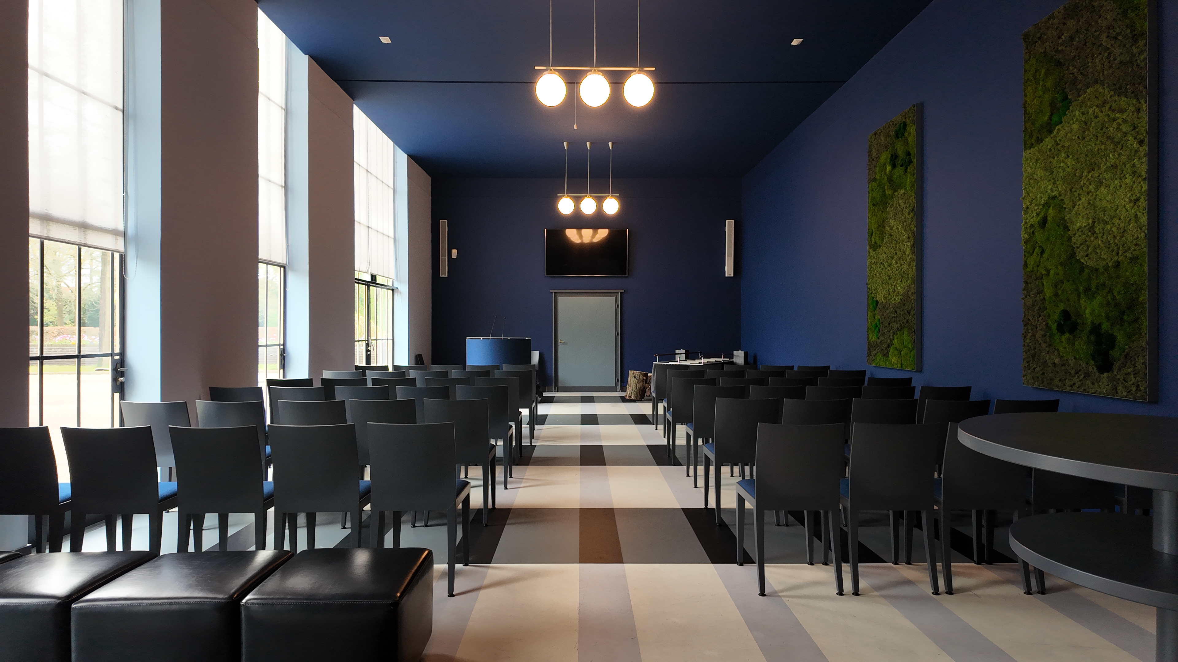Breed overzicht van het interieur van de aula met stoelen, ramen en blauwe wanden.