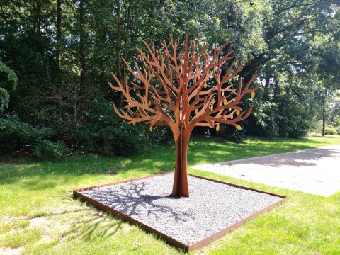 Metalen kunstwerk in de vorm van een boom, geplaatst op een lichte ondergrond in een groene omgeving.