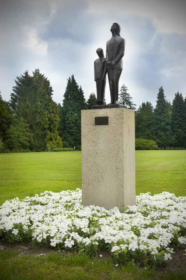 Standbeeld op een stenen sokkel, omringd door witte bloemen en bomen op de Noorderbegraafplaats in Hilversum.