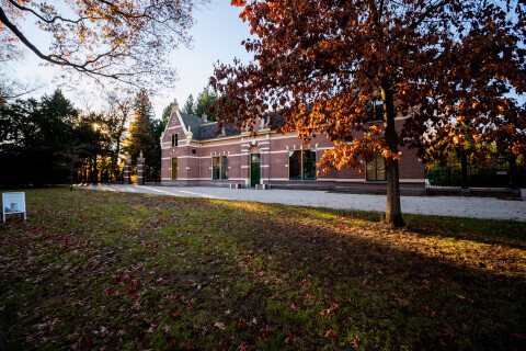 Historisch bakstenen gebouw op een groene begraafplaats, omgeven door bomen met herfstkleuren.