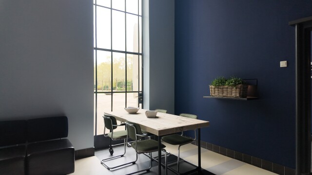 Kleine ontvangstruimte bij de aula met een tafel, hoge ramen en een blauwe wand.