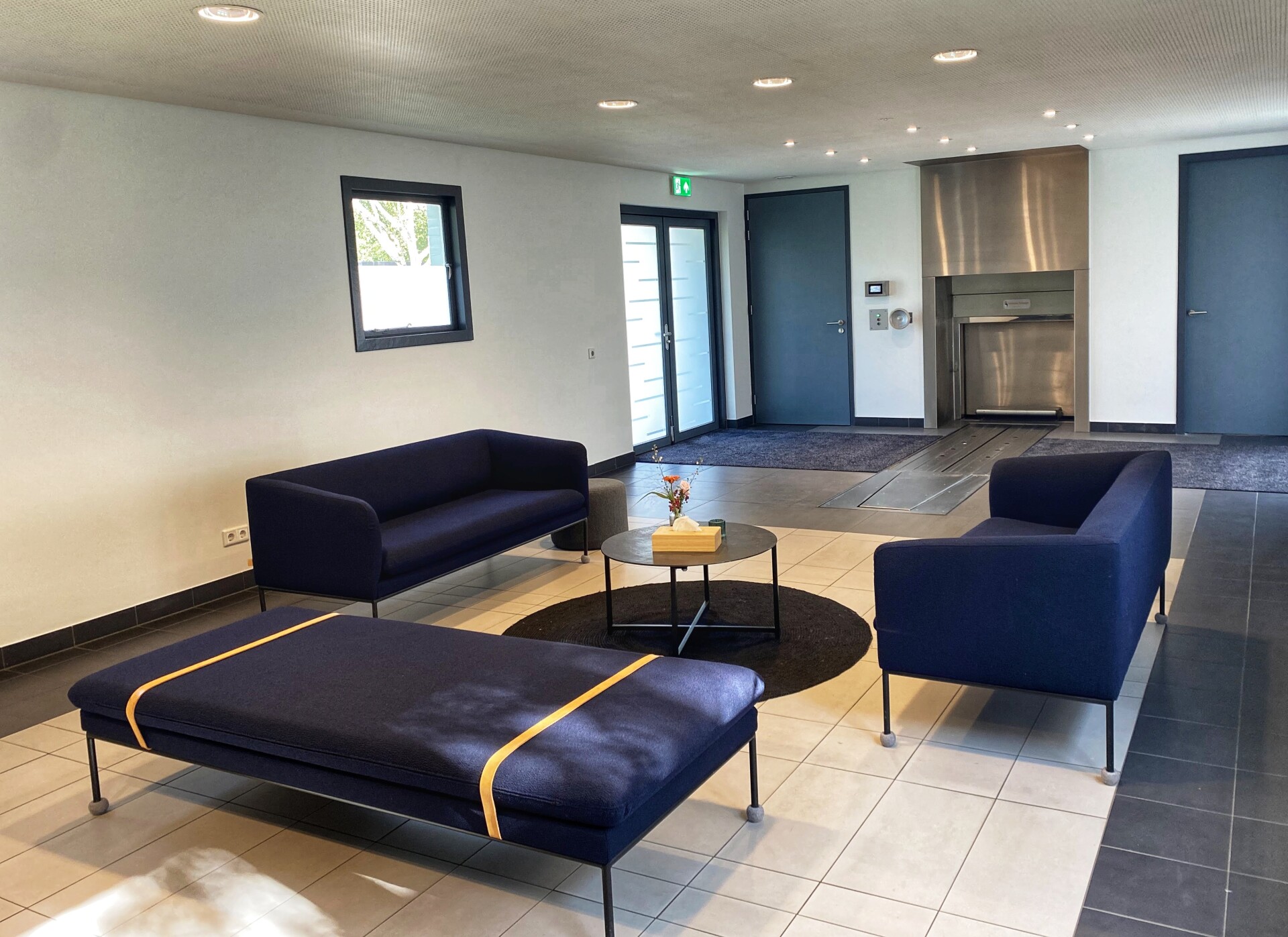 Ontvangstruimte van Crematorium Zuiderhof in Hilversum met blauwe zitbanken, een ronde tafel en een lichte inrichting.